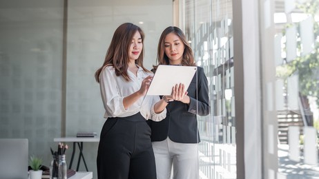 image of two young asian woman stand holding tablet by the window at the office.