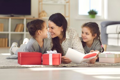 cute family celebrating mother's day at home. happy young woman lying on floor with her children, reading greeting card, thanking kids for present, touching noses and eskimo kissing with her daughters