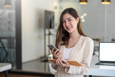 charming asian woman with a smile standing holding papers and mobile phone at the office.