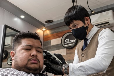an asian barber wearing a face mask trims and fades the side of a client's head with a hair clipper. a relaxed customer.