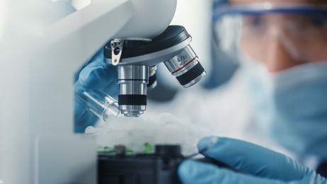 macro close up shot of male scientist in medical mask and safety glasses mixing green chemical liquid under the microscope. microbiologist working in modern laboratory with technological equipment.