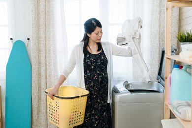 asian chinese pregnant woman adding clothes to washing machine and holding laundry basket in bright indoors. young maternity wife putting husband shirt inside. charming future mom doing housework.