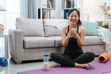 fit female athlete relax after training sit in sport yoga mat drinking from milk bottle. happy woman enjoy using mobile phone text message and resting after fitness workout. smiling girl on floor.