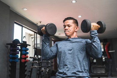 an asian man in a sweatshirt does seated arnold presses. shoulder workout and training. open air gym setting.