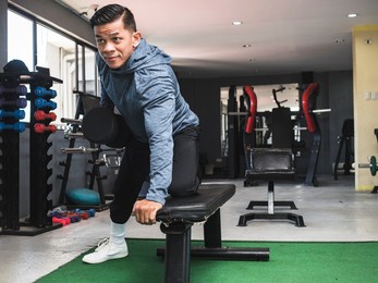 a fit asian man in a sweatshirt does one arm dumbbell rows. back workout exercise. inside a gym.