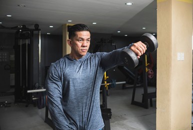 a fit asian man in a sweatshirt does alternating dumbbell front raises. shoulder workout and training. open air gym setting.