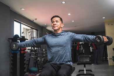 a fit asian man in a sweatshirt does seated side lateral raises. shoulder workout and training. open air gym setting.