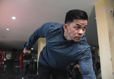 a determined asian man makes dumbbell triceps kickbacks on a bench at a gym. tricep and arm exercise and workout.