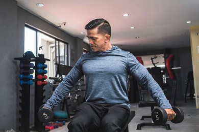 a fit asian man in a sweatshirt does seated side lateral raises. shoulder workout and training. open air gym setting.