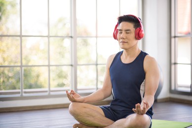 adult asian man wearing sport outfit using headphone to listen music or podcast and do exercise yoga workout for healthy self care at home