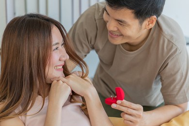 a young asian black hair male wearing brown shirt showing red diamond ring box in the left hand trying to propose marriage when asian long brown hair female smile exciting and sitting on orange sofa