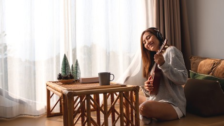 a beautiful young asian woman with headphone enjoy playing an ukulele at home