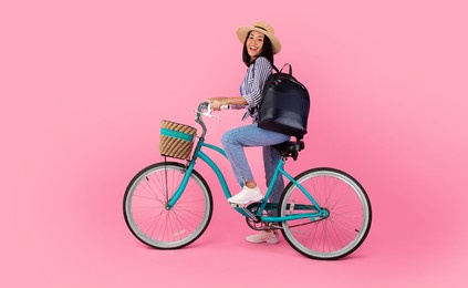 full length studio portrait of smiling young asian female cyclist wearing straw hat and black backpack posing on trendy retro bicycle with wicker basket isolated on pink background wall