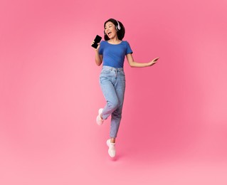 carefree singer. asian lady singing favorite song and using cell phone as a mic, dancing and jumping, wearing wireless headphones. excited woman having fun at pink studio, enjoying sound, full length