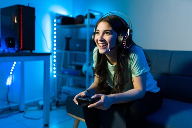 happy female gamer smiling while playing and winning in a video game with a remote controller. young woman sitting on the couch in her bedroom with neon led lights