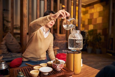 man pouring water into glass teapot while sitting at wooden table with cups and bowl of tea leaves