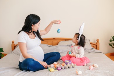 happy easter. asian chinese pregnant mother with baby girl playing with colorful easter eggs on bed at home. kid child and parent celebrating traditional christian holiday.
