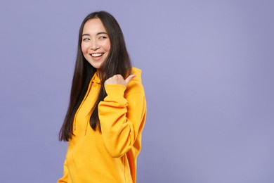 side view of smiling young brunette asian woman 20s wearing casual basic yellow hoodie standing pointing thumb aside on mock up copy space isolated on pastel violet colour background studio portrait