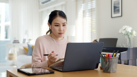 candid young attractive asian female employee wear earphone listen to digital online course development program in quarantine social distance at home reskill upskill school class for career path.