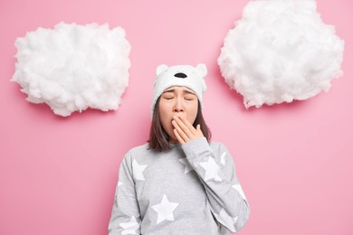 sleepy teenage asian girl covers mouth yawns wants to sleep has problem of insomnia dressed in nightwear soft bear hat isolated over pink background. bedtime energy charge concept. lack of vivacity