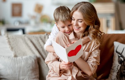 cheerful mother hugging son and reading handmade greeting card with heart while resting on sofa during holiday celebration mothers day at home