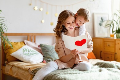 cheerful mother hugging son and reading handmade greeting card with heart while resting on bed during holiday celebration mothers day  at home