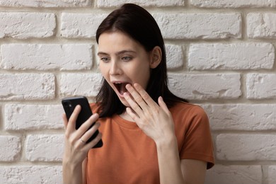 oh my god. amazed shocked female teenager looking on mobile phone screen with big eyes reading unexpected stunning news of getting big prize money reward winning lottery contest. brick wall background
