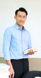 young asian male teacher holds a book and smiles at you in classroom