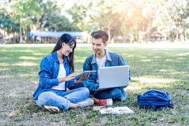 asian student couple studying outside on the grass, sitting down on the school campus using laptop computer, books, pencil and note book, studying hard under the trees nature in the evening sunlight