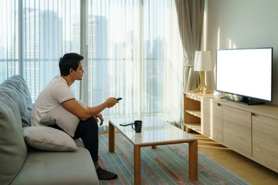an asian man holds a television remote and is pressing the channel while watching tv on the couch in the living room at home.