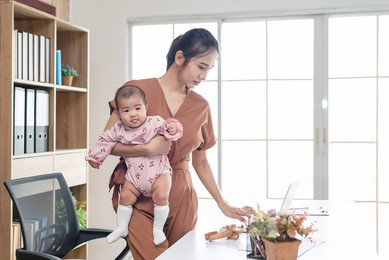 busy woman trying to work while babysitting newborn baby daughte.  young beautiful asian mother holding little baby on right hand while another hand typing and watching  business paper. working women 