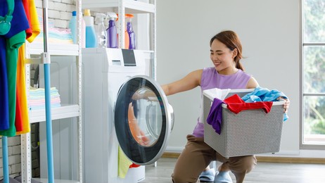 young happy beautiful friendly asian female long brown ponytail hair housekeeper wear purple sleeveless shirt and brown pants put dirty clothes from basket into white washing machine in laundry room.