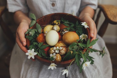 easter eggs and spring flowers in wooden bowl in hands on background of woman in rustic linen dress. stylish easter and quail eggs in natural dye and spring blooms. aesthetic holiday