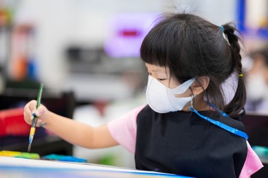 pupil wearing 3d medical face mask is sits in watercolor painting art in class. cute girl making crafts on canvas. child wear black apron. children concentrate on coloring work. kids aged 4 years old.