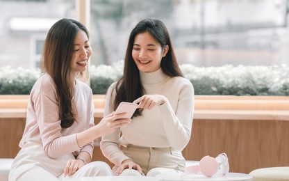 two young beautiful asian woman is using mobile phone and talking while sitting in coffee shop. technology and lifestyle concept.