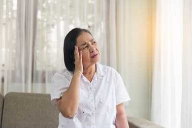 elderly asian woman sitting and having a headache and touching her head with her hands at home. retirement, quarantine and health care concept