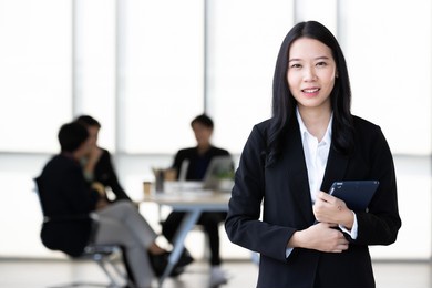 young asian businesswoman in black suit standing and holding tablet  computer posing to camera in office with her team talking in blur background.