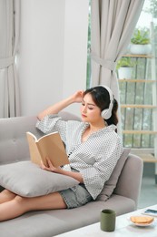 attractive asian young woman relax resting using smart phone for play music streaming application during read book sitting on couch or sofa at indoor garden home