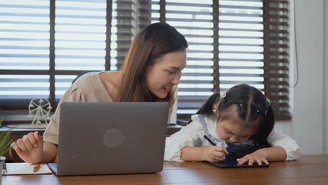 asian young mother working with laptop computer at home and help daughter doing homework, single mom freelancer work from home and helping his girl kid teaching homework on desk