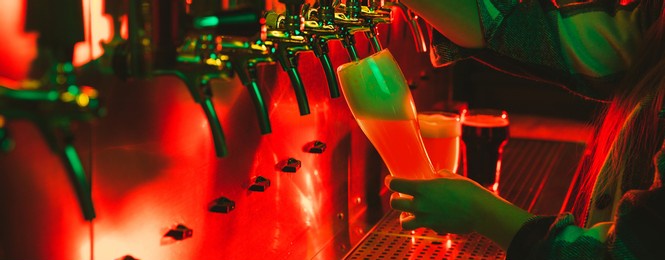 young woman barman pouring a cold lager beer from tap to glass in neon light.