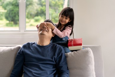 cute asian little daughter is closing father's eyes to surprise and give the present to daddy for birthday celebration, concept of love, relation and bonding of daddy and children in family lifestyle.