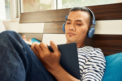 serious young asian man in headphones lying on bed, listening to music and reading book