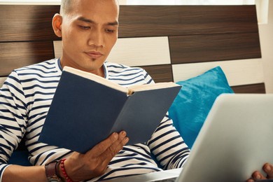 asian student sitting on bed, working on laptop and reading interesting book