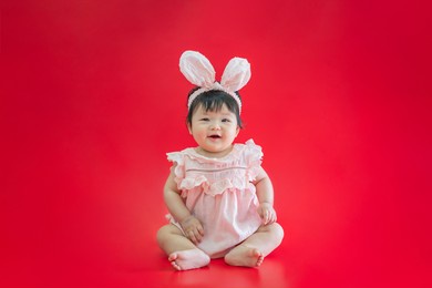 baby in easter bunny hairband. asian happy baby smiling and sitting on red background. cute 6 months baby start sitting with copy space as easter concept, baby or kid department in hospital.