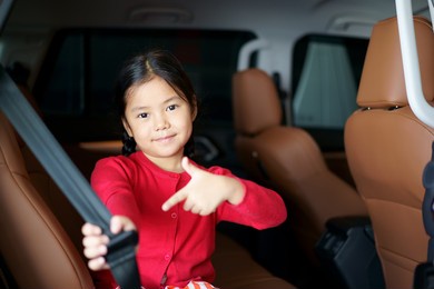 asian child cute or kid girl smile sit and show with point seatbelt or safety belt on brown back seat in car for safe in vehicle for transport holiday travel and wearing red shirt
