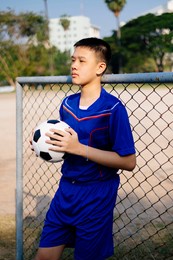 a boy in blue football suit standing and hold his ball at the fence.