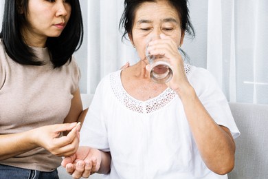 asian daughter taking care of old mother hand giving pills and glass of water helping her mom taking medicine on time 