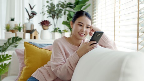 young attractive beautiful casual asia female lady student look at cellphone play social media sit at sofa couch living room feel relax comfort carefree with green houseplant at cozy home apartment.