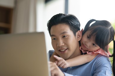 asian father on blue shirt working from home with his little cute girl daughter. asian kid hug his dad from behind while her father is working on a laptop.