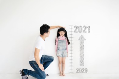 dad measures height growth of her child daughter at white brick wall with number date year 2020 to 2021